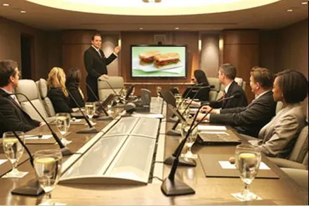 A man standing to present to a dozen colleagues seated around a long boardroom table set with microphones.