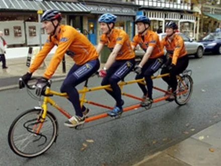 Four cyclists in matching orange kit riding a single long tandem bicycle down a street.